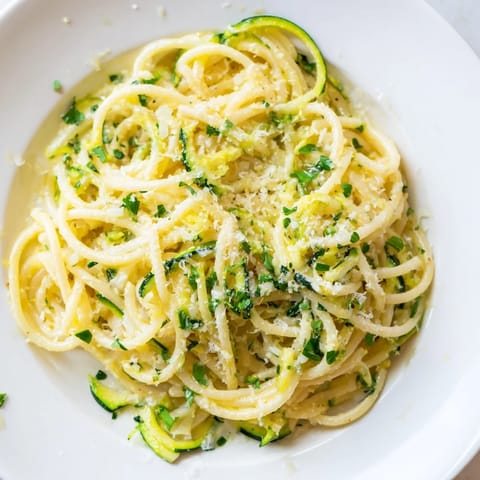 A close-up view shows steam rising from Lemon Zucchini Pasta, highlighting golden Parmesan shavings and a vibrant lemon wedge beside a colorful serving.  