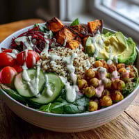 A vibrant Buddha Bowl with Quinoa, Roasted Sweet Potatoes, and Crispy Chickpeas is topped with fresh veggies and creamy garlic tahini dressing.
